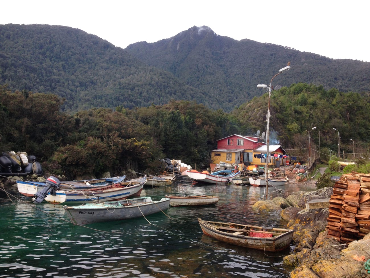 Boats moored in small harbour. House at edge of water, steep wooded hills behind.