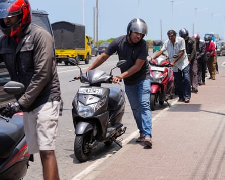 People in a queue push their scooters as they wait to refuel