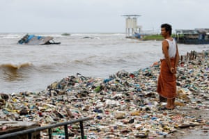 A villager stands among plastic and domestic waste on a beach as high tides sweep it to shore