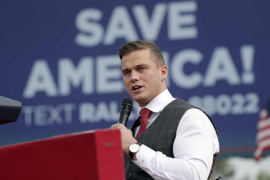 A man in a dress shirt, vest and tie speaks into a microphone. A sign behind him says 'Save America!'