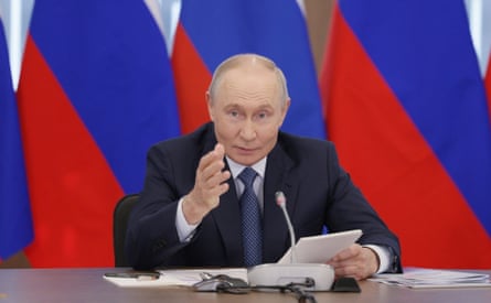 Vladimir Putin, sitting behind a table or desk, holds some papers in one hand and gestures with the other. Behind him are Russian flags