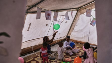 Four children of different ages mill around a woman who is sitting on the ground in a tent, gently batting a green balloon into the air. Above her are strings on which childrens’ drawings and pegged.
