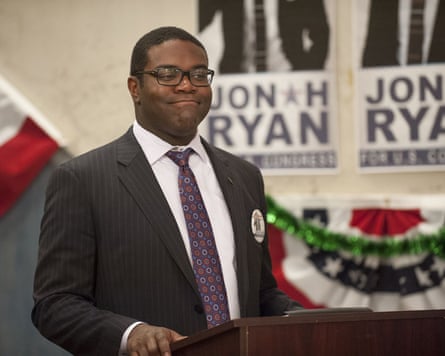 Man in suit at podium with flags and posters behind him
