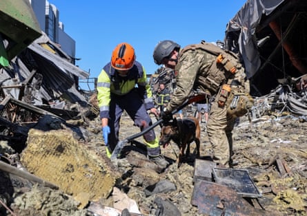 One rescue worker points to a spot in wreckage from the attack as another uses a shovel, with a dog standing next to them