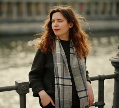 Naoise Dolan in a scarf, leaning against railings, a river behind her.