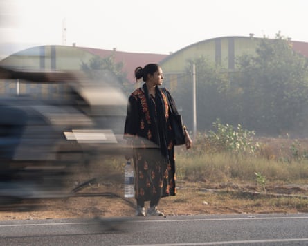 Neha looks to her left as she stands at the edge of a road while the blurred figure of a motorcylist moves past