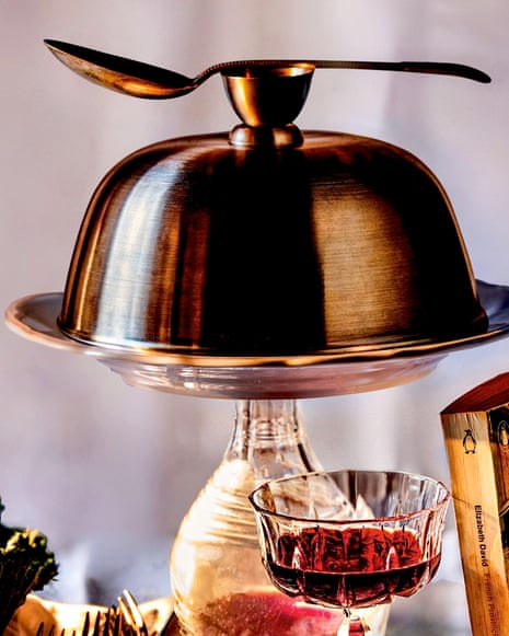 A domed metal serving dish (a cloche) with a spoon on top sits above a wine glass, decanter, cabbage, cutlery, and a cookbook on a patterned tablecloth.