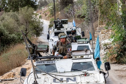 White armoured vehicles with UN and Unifil identification drive in convoy on a narrow road through greenery and a dry hillside; the vehicles carry blue UN flags and men with guns