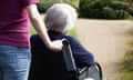 A woman holds the handles of a wheelchair in which an elderly woman with grey hair in sitting. They are facing away from the camera and neither of their faces are visible.