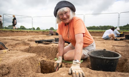 A woman in an orange t shirt and dark hate kneels on a sandy ground