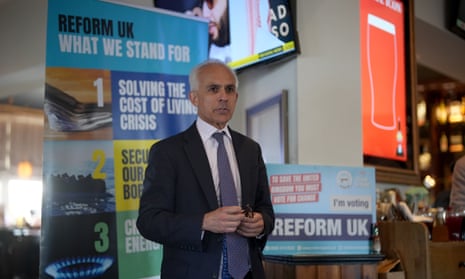 Ben Habib wearing a suit in front of a Reform UK campaign poster