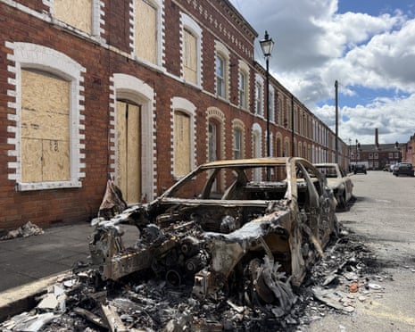 A burned-out car and boarded-up houses in a residential street