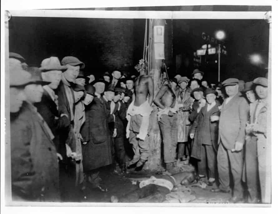 A crowd surrounds two African American men hanging from nooses on a pole.