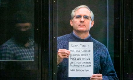 Paul Whelan, standing behind a glass panel, holds a sign aloft reading ‘Sham trial!’.