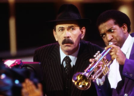 A man in a business suit and fedora hat with full black moustache bending to listen to a trumpet player in a purple suit