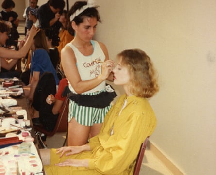 Bobbi Brown in the early days of her career, wearing green striped shorts and a vest, standing and putting makeup on a woman who is sitting