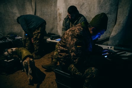 Soldiers look at handheld screens. A dog in a green coat is also in the picture