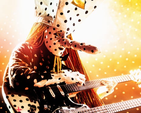 A person in a polka dot outfit and crown plays an electric guitar surrounded by falling confetti.
