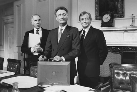 Nigel Hawthorne, Paul Eddington holding a briefcase and Derek Fowlds all in suits stand in front of a fireplace