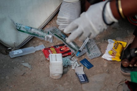 A pair of hands in latex gloves point to syringes, string, blades and pain relief packages on the ground. 
