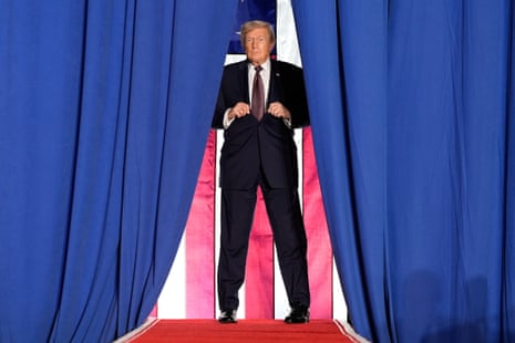 a man in a suit stands on a red carpet between a blue curtain