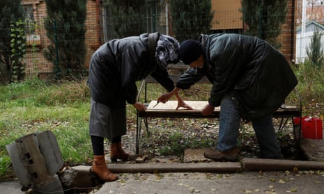 People repair the seat of a bench, as they stand next to an unexploded Smerch rocket after coming out of their underground shelters to receive aid.