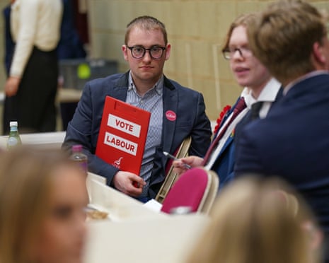 A man in glasses and a suit holds a red 'VOTE LABOUR' sign while seated at a vote count