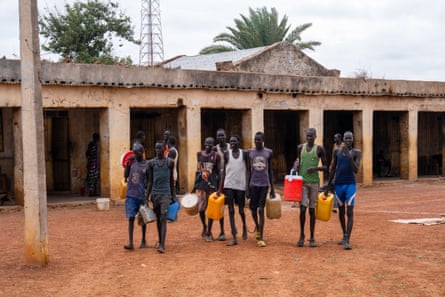 A group of young men walk through a dusty prison courtyard carrying jerrycans. They are chained in pairs at the ankle.