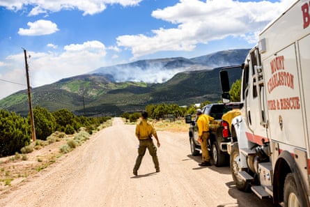 Firefighters stand in the road beside their truck as smoke rises from a hill in the distance