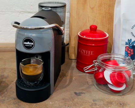 A grey coffee machine on a sideboard next to a cup of coffee, a jar of capsules and a red pot
