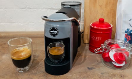 A grey coffee machine on a sideboard next to a cup of coffee, a jar of capsules and a red pot