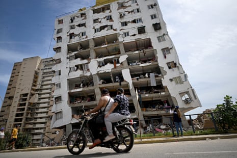 Two people ride a motorbike past a heavily damaged apartment building.