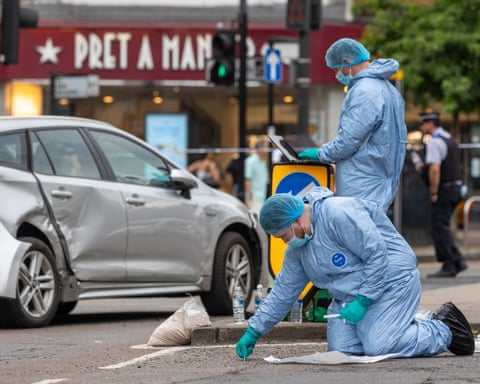 Two figures in blue protective clothing examine a road surface with a damaged silver car visible in the background.