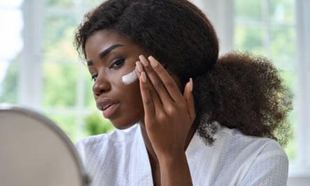 Black young woman applying cream on face skin looking in mirror.