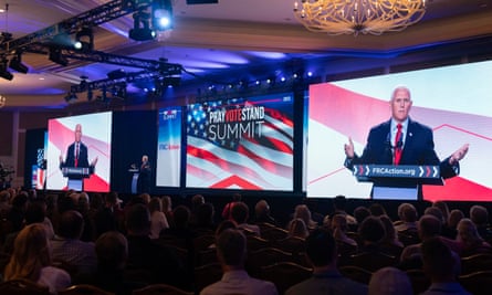 Taken from behind the darkened, seated crowd, we see them facing three mega-screens with images of an older, white-haired man in a suit and American flags, and much smaller, to the left, is the man actually on stage.