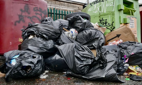 piles of black bin bags overflowing with rubbish against large waste and recycling containers