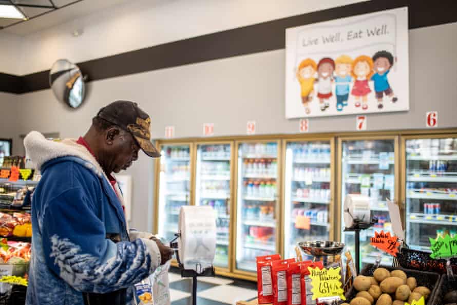 An elderly man wearing a Vietnam veterans hat stands in front of a bag dispenser in a grocery store trying to open a thin plastic produce bag. A display of potatoes is in front of him. In the background a refrigerated case can be seen.