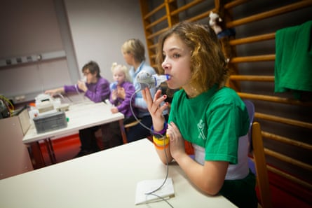 A young girl sits at a table holding an inhalation device to her mouth. Behins her two younger children can be seen sitting with a medical machine and an adult.