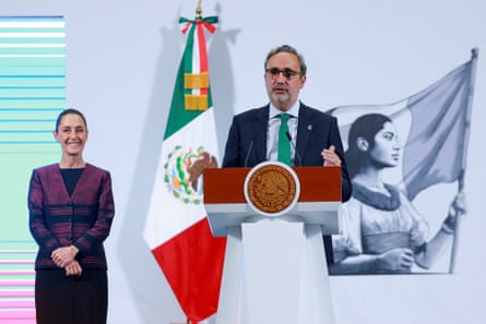 A man speaks at a podium with the Mexican flag behind him. A woman stands next to him
