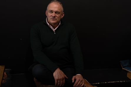 Ed Davey – portrait as he sits at home in a darkened room with his face and hands illuminated, facing the camera directly