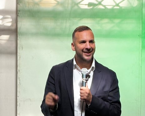 Green Party leader Zack Polanski holding a microphone gestures while speaking in front of a green backdrop