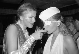 Black-and-white photo. A woman with a string of gemstones round her face holds a glass up to the mouth of another woman wearing an eyepatch