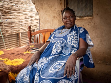 A middle-aged African woman sits in the porch of a house with mud walls