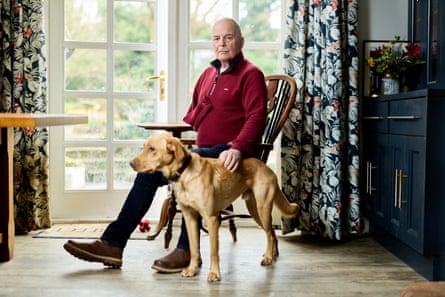 David Bulteel wearing a red sweat top with black trim, sitting in a chair beside a labrador-dog in front of a large french window with floral curtains.