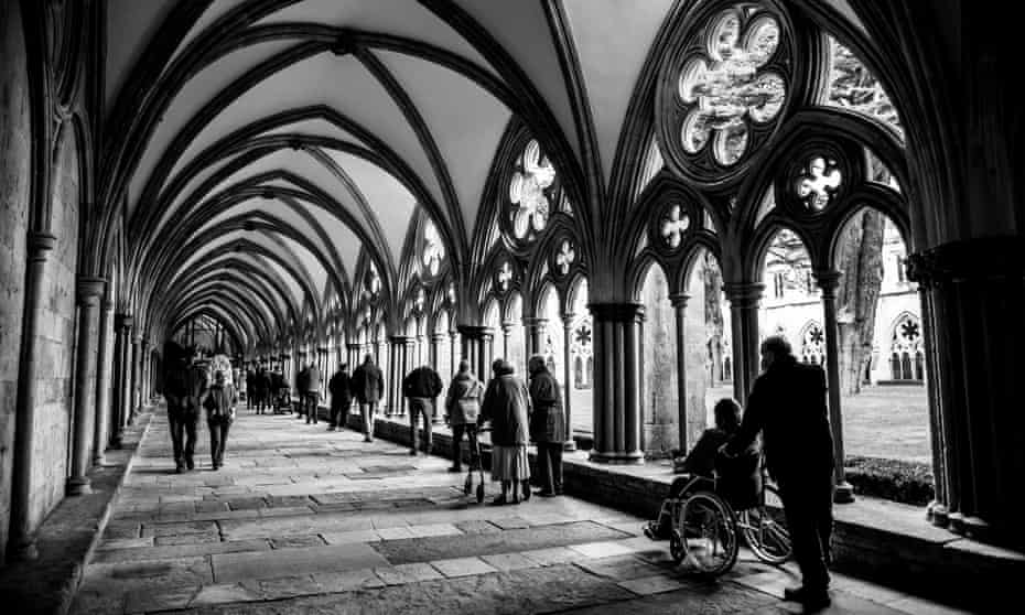 A high-contrast black and white photo looking down a long cloister at the cathedral, with a queue of socially distanced people waiting