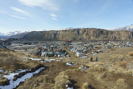 A lone hiker approaches a small town at the foot of a mountain. Patches of melted snow dot the landscape.