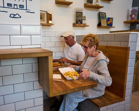 A man and a woman tuck into boxes of fish and chips at a wooden table in a white-tiled restaurant