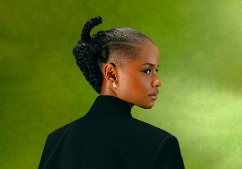 Letitia Wright in profile and photographed from behind, wearing a black coat and with her hair in elaborate plaits