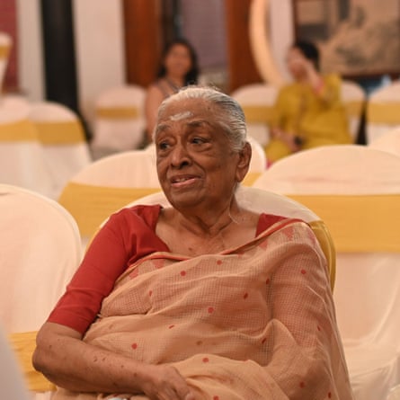 An older Indian woman wearing a peach-coloured sari sitting in a hall set up with rows of chairs