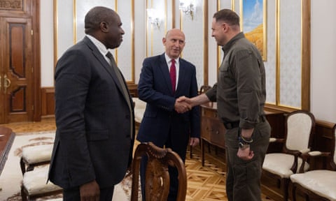 David Lammy stands to the side of John Healey as he shakes hands with Andriy Yermak; the three are standing in a ceremonial meeting room with decorative wooden parquet flooring, gold-framed panelling and dark wooden furniture. Lammy and Healey wear dark suits while Yermak is in military-style khakis. dqxikeidqkikdinv